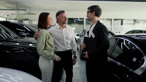 Happy-couple-a-middle-aged-man-with-gray-hair-together-with-his-brunette-wife-is-having-fun-talking-with-a-car-dealership-assistant-in-a-business-suit-about-cars-in-a-car-showroom