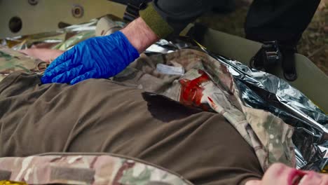 Close-up-a-confident-male-medic-in-a-dark-uniform-and-blue-rubber-gloves-uses-special-scissors-to-cut-the-T-shirt-of-a-soldier-wounded-in-the-chest-and-treats-the-wound-during-first-aid-training-in-combat-conditions