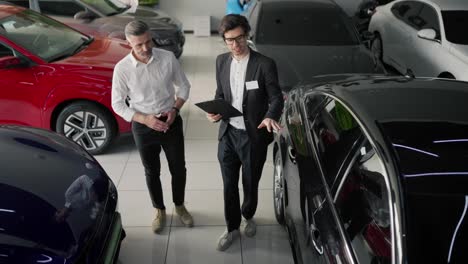 Top-view-of-a-confident-brunette-male-assistant-in-a-business-suit-talking-to-a-middle-aged-man-in-a-white-shirt-about-modern-car-models-and-talking-about-them-in-a-car-showroom