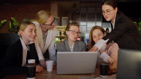 A-confident-middle-aged-blonde-girl-in-glasses-and-a-gray-uniform-communicates-with-her-businesswoman-colleagues-while-sitting-at-a-table-in-front-of-a-laptop-in-a-modern-office