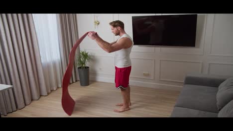 Happy-male-athlete-with-a-short-haircut-and-beard-at-home-in-a-sports-summer-suit-lays-out-a-mat-and-warms-up-before-playing-sports-inside-the-house.-Side-view-of-a-happy-blond-man-warming-up-in-a-white-t-shirt-and-red-shorts-before-starting-his-exercise-routine-at-home