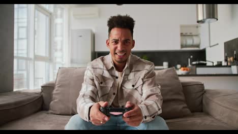 A-sad-and-angry-young-man-with-Black-skin-a-brunette-with-stubble-in-a-cream-colored-checkered-shirt-sits-on-a-sofa-in-a-modern-apartment-and-plays-a-game-using-a-black-joystick-and-is-sad-about-his-defeat-in-a-modern-apartment