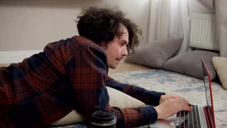 Confident-brunette-guy-with-curly-hair-in-a-checkered-shirt-writes-his-ideas-into-a-laptop-while-lying-on-the-floor-on-a-pillow-in-a-modern-apartment