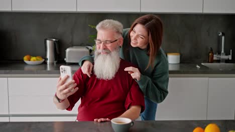 A-happy-and-cheerful-elderly-man-with-gray-hair-and-a-lush-beard,-together-with-his-adult-brunette-daughter-in-a-green-jacket-take-a-selfie-using-a-white-smartphone-in-a-modern-kitchen