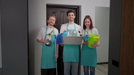 Portrait-of-a-happy-trio-of-cleaners-in-blue-fronts-with-various-tools-and-a-cleaner-for-cleaning-in-their-hands-two-men-and-a-brunette-girl-in-a-modern-apartment-on-a-cleaning-call