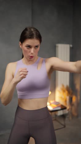 Vertical-video-of-a-confident-and-serious-brunette-girl-in-a-purple-sports-uniform-practicing-punches-with-her-hands-in-a-modern-apartment-at-home.-Confident-brunette-girl-in-a-purple-top-practices-boxing-punches-at-home-in-a-modern-apartment