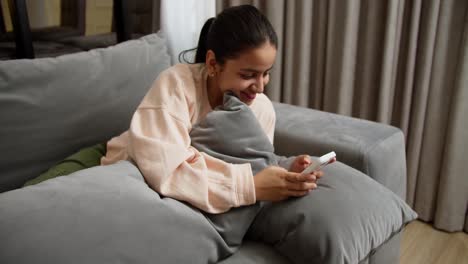 Happy-Indian-brunette-girl-in-a-light-sweater-and-green-pants-lies-on-the-sofa-hugging-a-pillow-and-typing-on-her-white-smartphone-while-relaxing-at-home