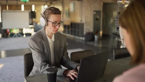 A-confident-blonde-girl-in-white-headphones-and-a-business-suit-sits-at-a-table-in-the-office-and-types-and-works-on-a-laptop.-A-middle-aged-woman-with-glasses-in-a-business-uniform-works-at-a-laptop-in-a-modern-office