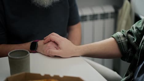 A-middle-aged-man-in-a-plaid-shirt-holds-hands-with-his-elderly-boyfriend-with-a-beard-while-sitting-at-a-table-in-the-kitchen-while-talking-about-their-relationship-as-an-LGBT-couple