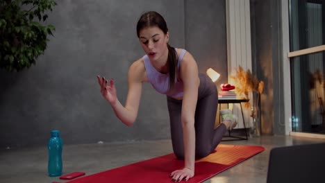 Confident-athletic-brunette-girl-in-a-purple-top-and-gray-pants-is-doing-sports-on-a-red-mat-using-video-lessons-using-laptops-in-a-modern-apartment-at-home