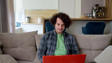 Happy-brunette-guy-in-a-checkered-shirt-with-curly-hair-rejoices-at-his-victory-while-working-using-a-laptop-on-the-sofa-in-a-modern-apartment