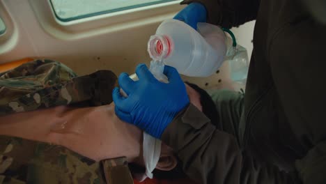 Close-up-of-a-confident-male-medic-in-a-dark-green-army-uniform-and-blue-gloves-performing-artificial-respiration-using-a-male-soldier-doll-during-first-aid-training-in-the-ambulance-cabin