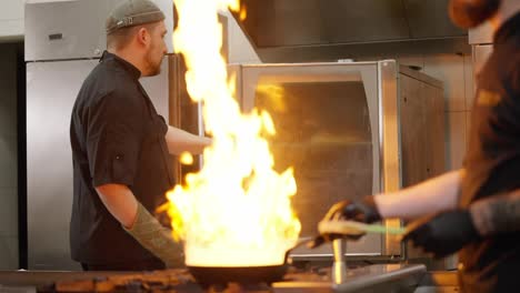 Aktive-Arbeit-In-Der-Küche-Eines-Restaurants-Ein-Koch-Zündet-Mit-Alkohol-Ein-Feuer-In-Einer-Bratpfanne-An,-Während-Sein-Kollege-In-Der-Küche-Eines-Restaurants-Die-Fertigen-Backwaren-Aus-Dem-Ofen-Nimmt