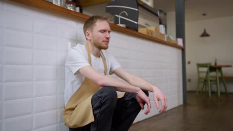 A-tired-blond-male-waiter-in-a-yellow-apron-rubs-his-head-and-sits-near-a-small-wall-in-a-cafe