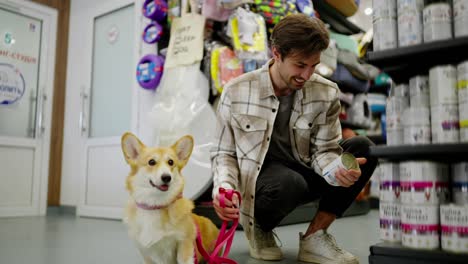 Confident-brunette-guy-in-a-light-checkered-shirt-chooses-food-for-his-yellow-corgi-dog-in-a-pet-store.-Confident-brunette-guy-with-his-dog-in-a-pet-store