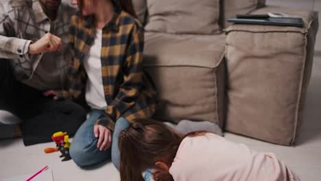 A-little-brunette-girl-in-a-pink-suit-draws-on-the-floor-on-paper-while-her-parents-communicate-with-each-other-and-sit-near-the-girl-near-the-gray-brown-in-a-modern-studio-apartment