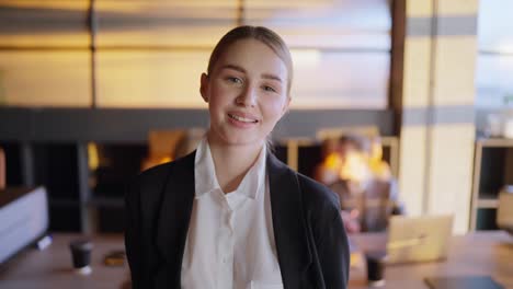 Portrait-of-a-happy-blonde-girl-in-a-black-business-uniform-posing-near-her-colleagues-from-the-table-in-a-sunny-office.-Confident-young-girl-businesswoman-posing-in-the-office