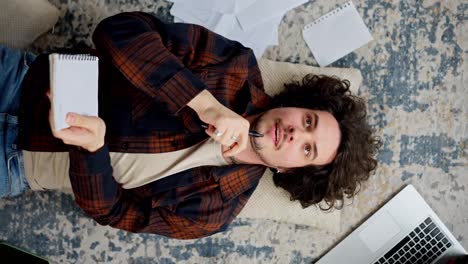 A-thoughtful-brunette-guy-with-curly-hair-in-a-checkered-shirt-lies-on-the-floor-on-a-pillow-and-writes-down-his-ideas-in-a-white-notebook-among-many-papers-and-a-laptop-at-home