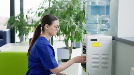 A-confident-and-tired-brunette-doctor-girl-in-round-glasses-and-a-blue-uniform-pours-water-from-a-cooler-into-a-paper-glass-and-drinks-fresh-water-during-a-break-at-work-in-a-modern-clinic