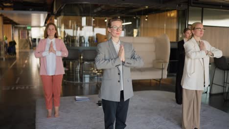 Group-of-confident-businesswomen-in-business-suits-doing-yoga-on-a-gray-soft-carpet-in-a-modern-office-during-a-break-at-work.-Yoga-classes-and-spiritual-support-while-working-in-the-office