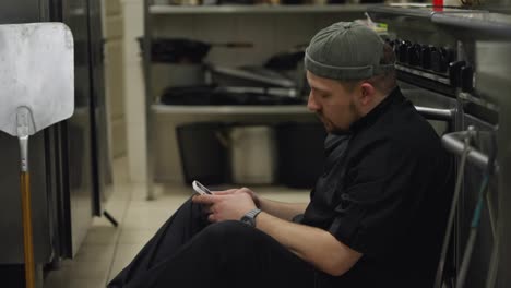 A-tired-cook-in-a-black-uniform-with-a-hat-sits-on-the-floor-in-the-kitchen-leaning-on-the-table-and-is-distracted-by-social-networks-on-his-smartphone-during-a-short-break-and-relaxation-on-a-hard-day-at-work-in-the-kitchen-of-a-restaurant