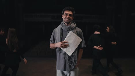 Portrait-of-a-happy-young-male-stage-director-with-papers-in-his-hands-with-a-scarf-and-T-shirt-on-stage-in-the-theater-during-a-rehearsal