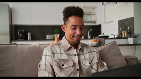 A-happy-young-Black-skinned-brunette-man-with-stubble-in-a-checkered-beige-shirt-sits-on-a-modern-gray-sofa-and-works-on-his-laptop-remotely-while-at-home-in-a-studio-apartment