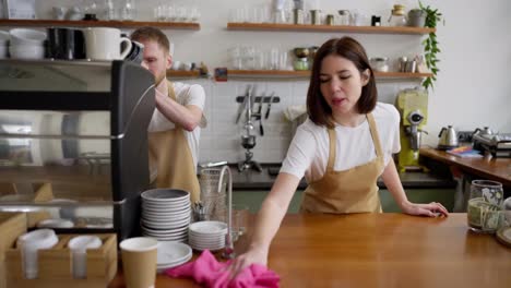 A-brunette-girl-in-a-yellow-apron-cleans-the-counter-with-a-pink-rag-while-preparing-for-a-working-day-with-a-barista-guy-in-a-cafe