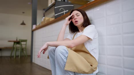 A-tired-girl-waitress-in-a-yellow-apron-sits-on-the-floor-near-the-cash-register-in-a-cafe-and-rubs-her-head-with-her-hands-during-a-hard-day-at-work