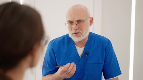 Por-Encima-De-Su-Hombro,-Un-Médico-Mayor-Y-Seguro-De-Sí-Mismo,-Con-Gafas-Y-Un-Uniforme-Azul-Con-Un-Pequeño-Micrófono-En-El-Pecho,-Se-Comunica-Y-Da-Una-Entrevista-A-Una-Joven-Morena-Con-Gafas-Sobre-Su-Trabajo-En-La-Nueva-Clínica-Moderna.