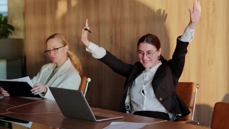 Happy-brunette-businesswoman-in-round-glasses-spins-on-a-chair-at-a-desk-in-the-office-while-fighting-for-success-and-victory-at-her-desk-in-a-modern-office