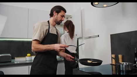 Happy-brunette-man-with-stubble-in-a-black-apron-and-beige-T-shirt-prepares-cheesecakes-during-breakfast-with-his-brunette-wife-in-a-modern-apartment.-Cooking-pancakes-and-preparing-for-breakfast-in-a-modern-kitchen