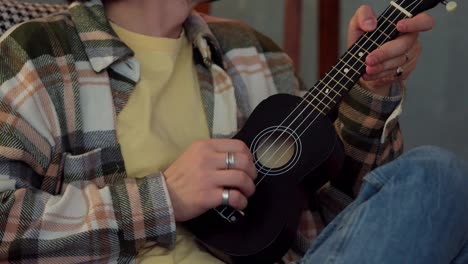 Close-up-of-a-guy-with-curly-brunette-hair-in-a-checkered-shirt-playing-the-ukulele-with-his-hand-with-two-rings-in-a-modern-apartment