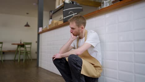 Tired-male-blond-waiter-in-a-yellow-apron-rubs-his-head-with-his-hands-and-exhales-during-a-hard-day-at-work