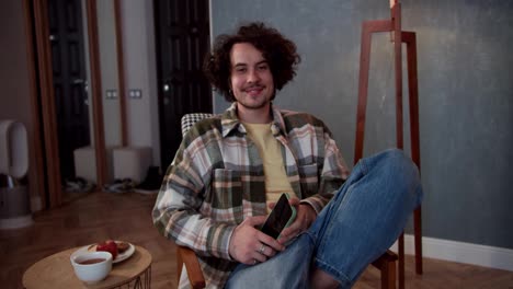 Portrait-of-a-happy-brunette-guy-with-curly-hair-in-a-checkered-shirt-who-holds-a-smartphone-in-his-hands-and-sits-on-a-comfortable-chair-in-a-modern-cozy-apartment