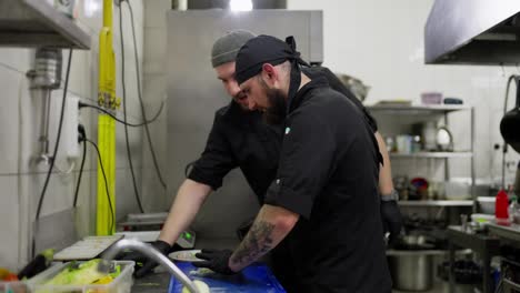 Two-professional-chefs-in-black-uniforms-work-on-preparations-and-cut-vegetables-while-working-in-a-restaurant-kitchen.-A-male-cook-chops-an-onion-while-his-colleague-takes-the-chopped-and-prepared-vegetables-putting-them-into-the-refrigerator-in-the-kitchen