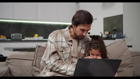 Ein-Brünetter-Mann-Mit-Stoppeln-Und-Beigefarbenem-Hemd-Kommuniziert-Mit-Seiner-Kleinen-Tochter,-Während-Er-Arbeitet-Und-Zeit-An-Einem-Grauen-Laptop-Auf-Dem-Sofa-In-Einem-Modernen-Apartment-Verbringt