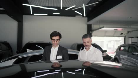 A-confident-brunette-guy-with-glasses-and-a-business-suit-an-assistant-at-a-car-showroom-tells-a-middle-aged-man-in-a-white-shirt-about-modern-cars-at-the-car-showroom