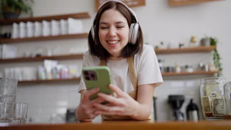 Close-up-of-a-happy-brunette-girl-waiter-in-white-headphones-listens-to-music-and-plays-a-video-on-a-green-smartphone-standing-at-the-counter-in-a-cafe