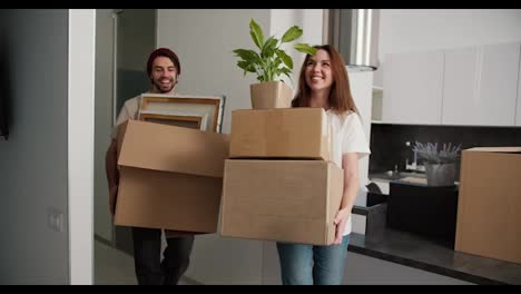 Ein-Glückliches-Paar,-Ein-Brünettes-Mädchen-In-Einem-Weißen-T-Shirt-Und-Ihr-Brünetter-Freund-Mit-Stoppeln-In-Einem-Cremefarbenen-T-Shirt-Bringen-Kisten-Mit-Sachen-In-Ihre-Neue-Moderne-Wohnung-Und-Stellen-Die-Kisten-Auf-Das-Sofa,-Das-Mit-Einer-Plastikhülle-Bedeckt-Ist.