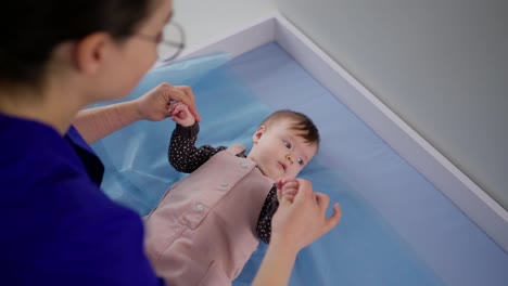 From-above-over-the-shoulder-a-confident-brunette-girl-doctor-in-glasses-finds-a-common-language-and-checks-a-little-baby-girl-in-a-pink-overalls-for-the-correct-development-in-a-modern-clinic-at-a-pediatricians-appointment