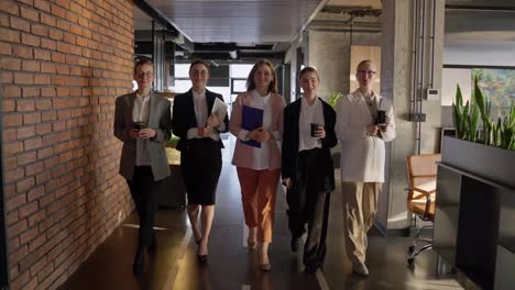 Portrait-of-a-group-of-five-confident-businesswomen-in-business-clothes-with-coffee-and-laptops-in-their-hands-walking-along-a-gray-floor-in-a-modern-office