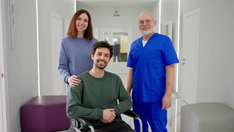 Portrait-of-a-Happy-brunette-man-with-stubble-in-a-Green-jacket-who-sits-on-a-wheelchair-after-being-injured-near-a-brunette-girl-and-an-experienced-confident-male-doctor-in-a-blue-uniform-in-a-modern-clinic