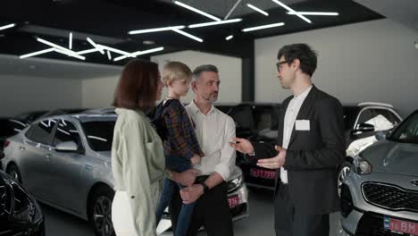 Ein-Selbstbewusster-Junger-Brünetter-Mann-In-Einem-Business-Anzug-Und-Einer-Brille,-Ein-Assistent-In-Einem-Autohaus,-Erklärt-Einem-Mann-Seiner-Frau-Und-Seinem-Kleinen-Sohn,-Was-Autos-Sind-Und-über-Sie