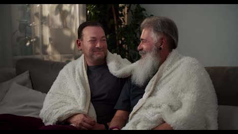 A-happy-brunette-man-in-a-gray-T-shirt-wrapped-in-a-white-woolen-blanket-sits-on-the-sofa-and-talks-with-his-elderly-boyfriend-with-gray-hair-and-a-lush-white-beard-in-a-modern-apartment.-LGBT-couple-communicates-and-spends-time-together