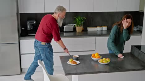 An-elderly-man-with-gray-hair-and-a-beard-in-a-red-T-shirt-is-cleaning-the-kitchen-with-a-mop-along-with-his-adult-brunette-daughter-in-a-green-sweater-in-a-modern-kitchen