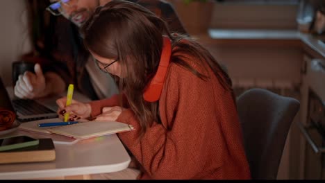 A-young-confident-brunette-girl-in-red-wireless-headphones-and-an-orange-sweater-studies-foreign-languages-with-her-teacher-Brunet-and-takes-notes-in-her-notes-while-studying-foreign-languages-in-a-modern-apartment-in-the-kitchen