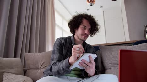 Focused-brunette-guy-with-curly-hair-and-mustache-in-a-gray-checkered-shirt-takes-notes-and-writes-down-important-points-while-watching-an-educational-video-on-a-red-laptop-while-sitting-on-the-couch-at-home