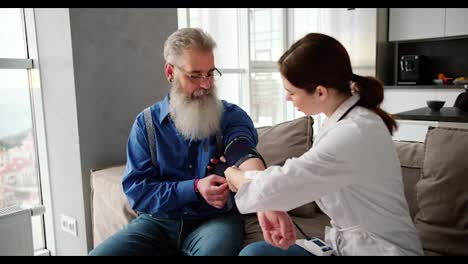 Una-Doctora-Morena-Con-Una-Bata-Blanca-Examina-A-Un-Hombre-Mayor-Con-Cabello-Gris-Y-Una-Barba-Exuberante-Con-Una-Camisa-Azul-Usando-Un-Monitor-De-Frecuencia-Cardíaca-Y-Mide-La-Presión-Arterial-Del-Hombre-En-Un-Apartamento-Moderno.