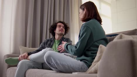 Happy-brunette-guy-with-curly-hair-in-a-gray-checkered-shirt-communicates-with-a-brunette-girl-while-sitting-on-the-sofa-at-home